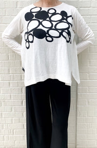 Front top half view of a woman wearing the moyuru long sleeve circle top. This top is white with black filled and empty circles on the top half. The top has fitted sleeves and a flowy body.