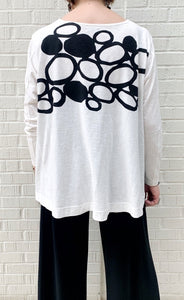 Back top half view of a woman wearing the moyuru long sleeve circle top. This top is white with black filled and empty circles on the top half. The top has fitted sleeves and a flowy body.