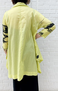 Back full body view of a woman wearing the moyuru art print tunic. This tunic is yellow with black abstract print on the sleeves, a solid back, and a hem that sits below the hips at the knees.