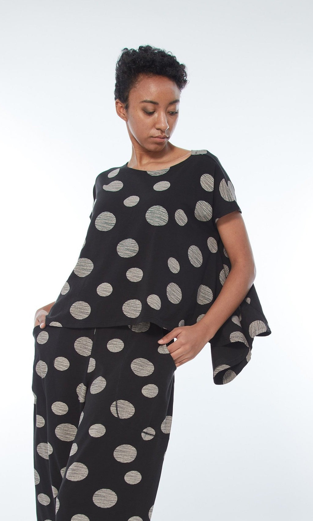 Front top half view of a woman wearing the M x Matthildur Ashley top. This top is black with taupe dots on it. The top has short sleeves and a flowy fit with an asymmetrical hem. On the bottom the model is wearing a matching black and taupe dotted pant.