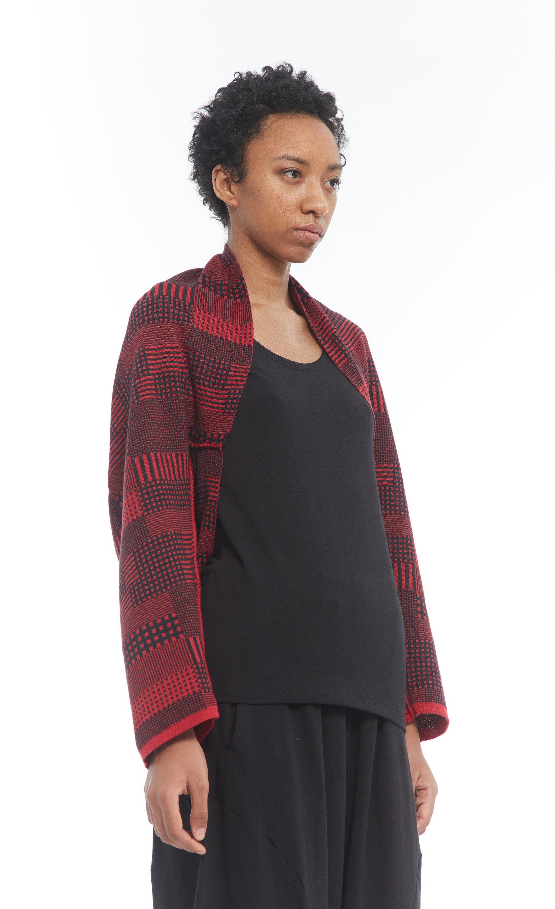 Front top half view of a woman wearing the mxmatthildur vonji sweater. This sweater is red and black plaid patchwork. The sweater has long sleeves and is being worn as a cropped shrug with a black tank.