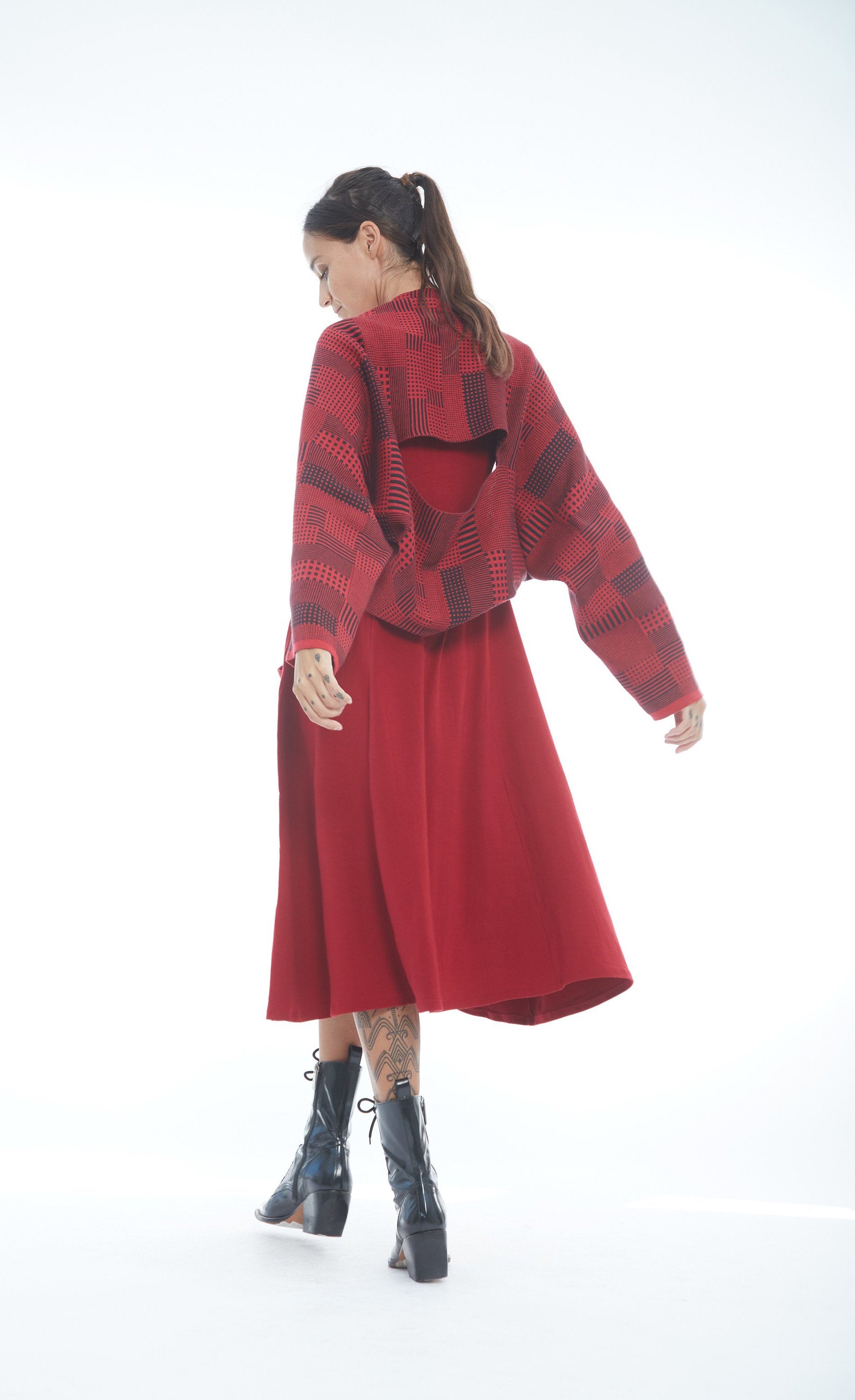 Back full body view of a woman wearing the mxmatthildur vonji sweater. This sweater is red and black plaid patchwork. The sweater has long sleeves and is being worn as a cropped shrug with a red dress.