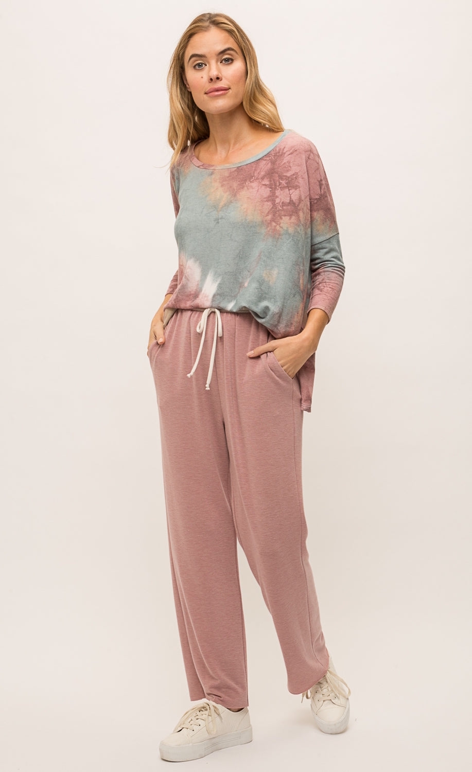 Front full body view of a woman wearing the mystree vintage tie dye top and the mystree lounge pant. The top is pink and blue tie dye and has long drop shoulder sleeves and a boxy fit. The bottoms are burgundy pink with a relaxed fit, pockets, and a white waistband.