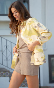 Side view of a woman wearing a matching ribbed and taupe colored tank and short. Over the tank she is wearing the Mystree Camo Jacket. This jacket is different shades of yellow camo with some brown mixed in. The front has buttons and a two large pockets. 
