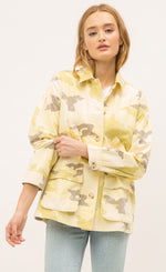Load image into Gallery viewer, Front view of a woman pulling up the sleeve of the Mystree Camo Jacket she is wearing. This jacket is different shades of yellow camo with some brown mixed in. The front has buttons and a two large pockets. 
