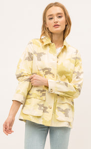 Front view of a woman pulling up the sleeve of the Mystree Camo Jacket she is wearing. This jacket is different shades of yellow camo with some brown mixed in. The front has buttons and a two large pockets. 