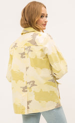 Load image into Gallery viewer, Back top half view of a woman wearing the Mystree Camo Jacket. This jacket is different shades of yellow camo with some brown mixed in. The back has a small slit near the middle of the bottom of the jacket.
