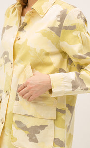 Close up front view of a woman with her thumb in the pocket of the Mystree Camo Jacket she is wearing. This jacket is different shades of yellow camo with some brown mixed in. The close up focuses on the jacket cuff and large, front left-sided pocket.