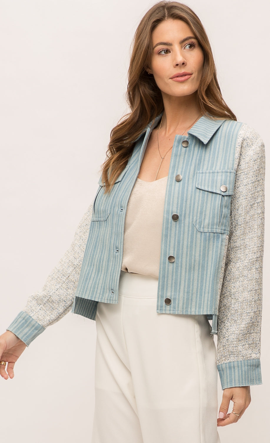 Front top half view of a woman wearing the Mystree Mix Media Jean Jacket with an off-white pant and over an off-white tank. This jacket has a striped denim body with woven grey and blue sleeves and sides. The front has two patch breast pockets and a button up front. 