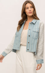 Load image into Gallery viewer, Front top half view of a woman wearing the Mystree Mix Media Jean Jacket with an off-white pant and over an off-white tank. This jacket has a striped denim body with woven grey and blue sleeves and sides. The front has two patch breast pockets and a button up front. 
