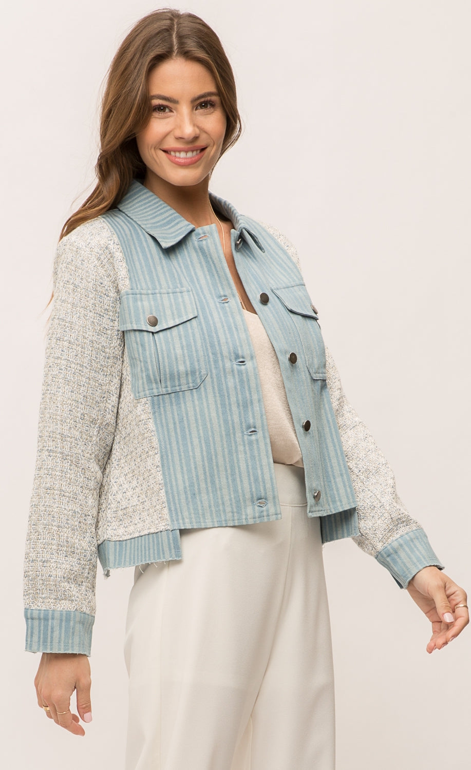 Front, right sided top half view of a woman wearing the Mystree Mix Media Jean Jacket with an off-white pant and over an off-white tank. This jacket has a striped denim body with woven grey and blue sleeves and sides. The front has two patch breast pockets and a button up front. 