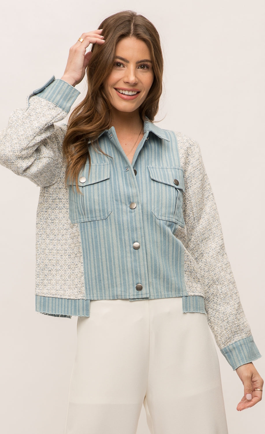 Front top half view of a woman wearing the Mystree Mixed Media Jean Jacket closed with an off-white pant. This jacket has a striped denim body with woven grey and blue sleeves and sides. The front has two patch breast pockets and a button up front. 