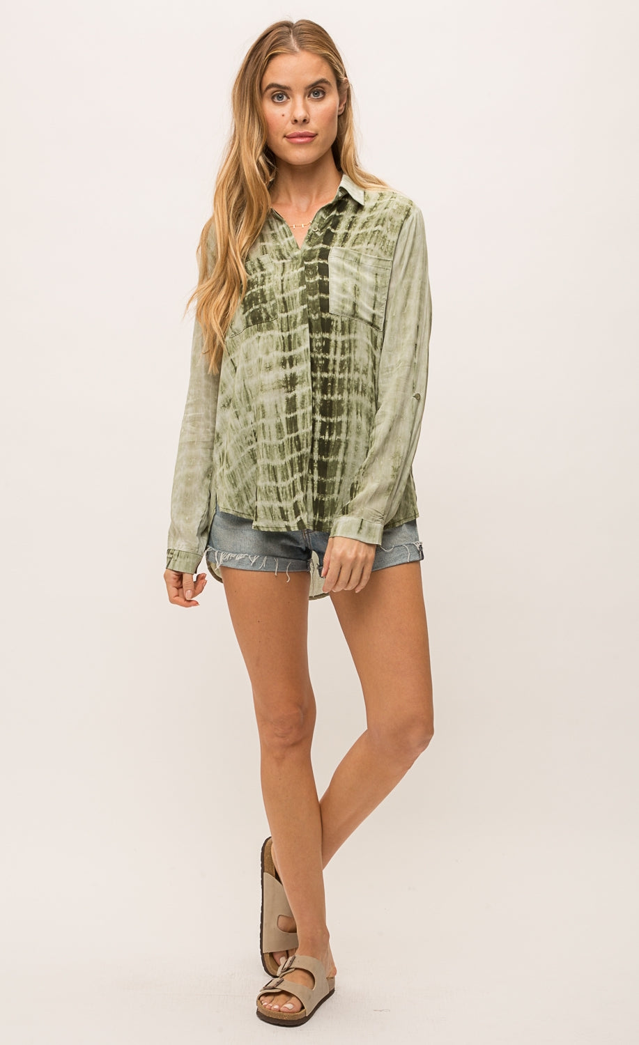 Front full body view of a woman wearing jean shorts and the Mystree Flowy Tie Dye Shirt. The shirt is olive green tie dyed with a hidden button up front, two breast pockets and long sleeves.