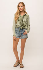 Load image into Gallery viewer, Front full body view of a woman wearing jean shorts and the Mystree Flowy Tie Dye Shirt tied in the front. The shirt is olive green tie dyed with a hidden button up front, two breast pockets and long sleeves.
