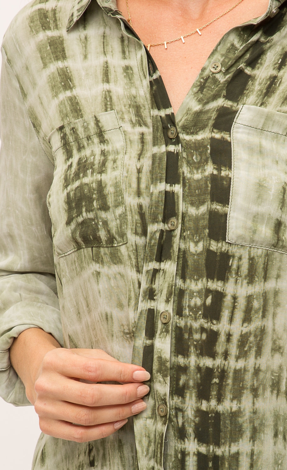 Front close up view of the hidden front buttons of the Mystree Flowy Tie Dye Shirt she is wearing. The shirt is olive green tie dyed with two breast pockets and long sleeves.