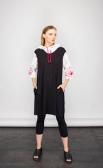 Load image into Gallery viewer, Front full body view of a woman wearing the NY 77 design flower sweatshirt tunic. This tunic looks like a hoodie layered under a sleeveless tunic but it is one piece. The tunic portion is black with front pockets. The hoodie portion is white with red flowers drawn on it and red drawstrings. The sleeves are 3/4 length.
