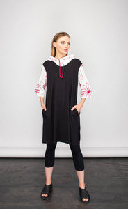 Front full body view of a woman wearing the NY 77 design flower sweatshirt tunic. This tunic looks like a hoodie layered under a sleeveless tunic but it is one piece. The tunic portion is black with front pockets. The hoodie portion is white with red flowers drawn on it and red drawstrings. The sleeves are 3/4 length.