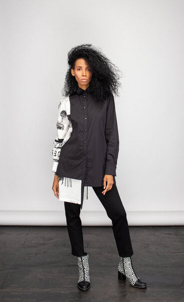 Front full body view of a woman wearing the ny 77 design mixed shirt. This shirt is predominately black. It has a button down front and long sleeves. The left side of the shirt is solid black. The right side of the shirt is white with abstract sketched print on it. The right side's hem is longer than the left.