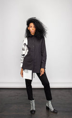 Load image into Gallery viewer, Front full body view of a woman wearing the ny 77 design mixed shirt. This shirt is predominately black. It has a button down front and long sleeves. The left side of the shirt is solid black. The right side of the shirt is white with abstract sketched print on it. The right side&#39;s hem is longer than the left.
