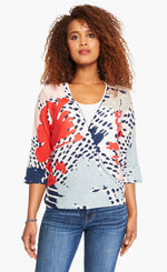 Load image into Gallery viewer, Front top half view of a woman wearing the nic+zoe art scene 4-way cardigan over a white tank. This cardigan has a multicolored red, blue, and tan print on a white base. The cardigan also has an open front and a pointed hem. In this image the cardigan wrapped to the side..
