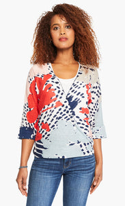 Front top half view of a woman wearing the nic+zoe art scene 4-way cardigan over a white tank. This cardigan has a multicolored red, blue, and tan print on a white base. The cardigan also has an open front and a pointed hem. In this image the cardigan wrapped to the side..