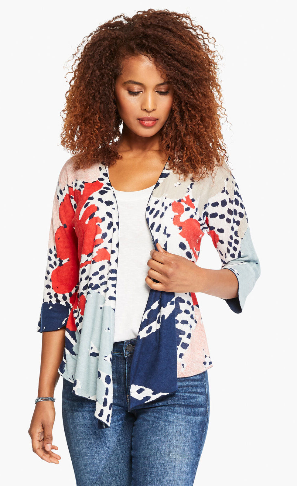 Front top half view of a woman wearing the nic+zoe art scene 4-way cardigan. This cardigan has a multicolored red, blue, and tan print on a white base. The cardigan also has an open front and a pointed hem. 