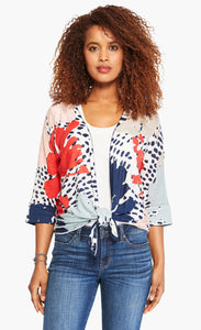 Front top half view of a woman wearing the nic+zoe art scene 4-way cardigan over a white tank. This cardigan has a multicolored red, blue, and tan print on a white base. The cardigan also has an open front and a pointed hem. In this image the cardigan is tied in the front.