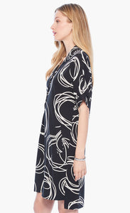 Left full body view of a woman wearing the nic+zoe billow dress. This dress is black with white swirl print all over it. The dress has a 3/4 button up front, elbow length sleeves, and front pockets. The dress sits at the knees.