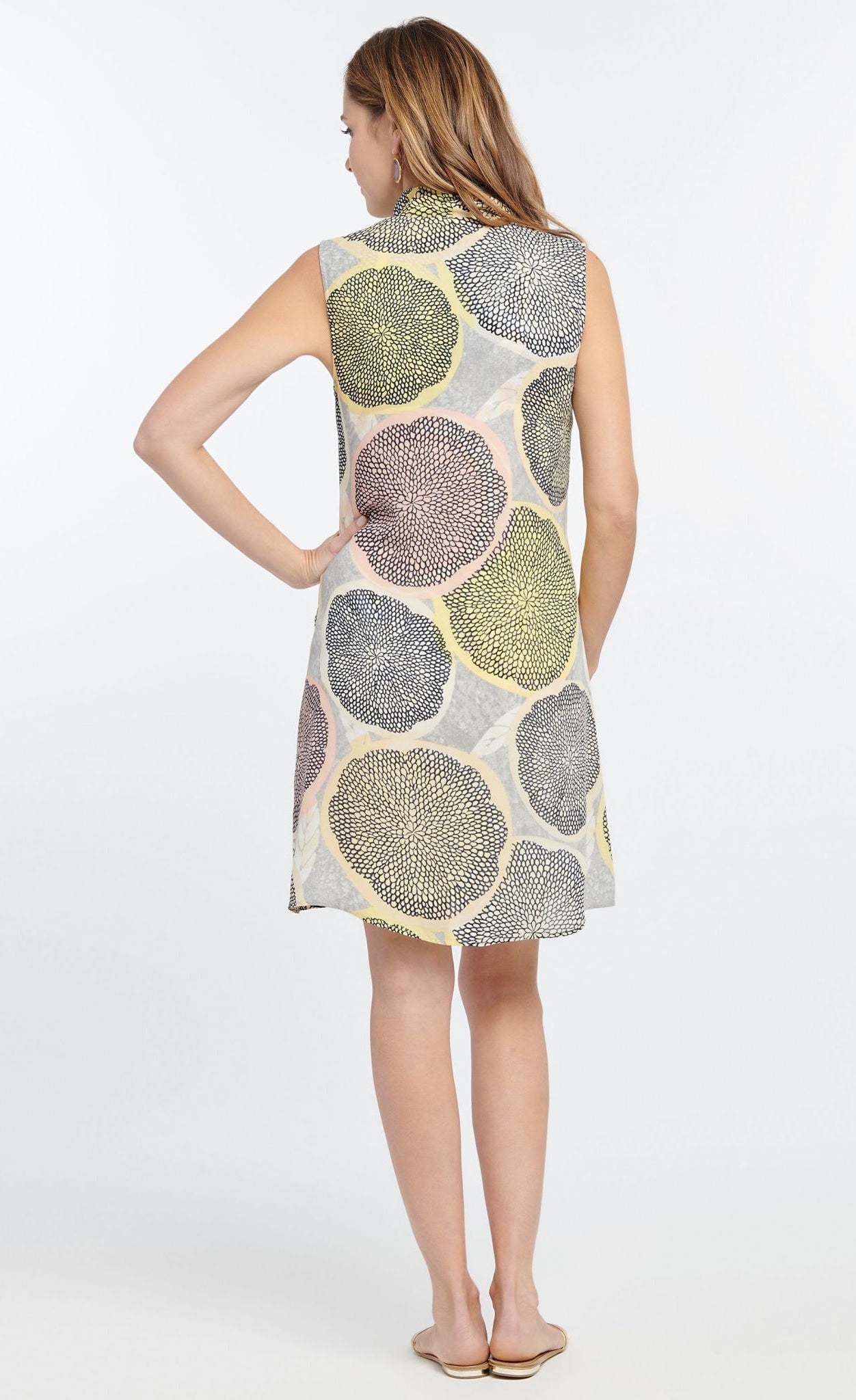Back full body view of a woman wearing the nic + zoe citrus dress. This dress is sleeveless and sits above the knees. It is pale yellow and grey with a large citrus slices print in yellow, blow and pink. 