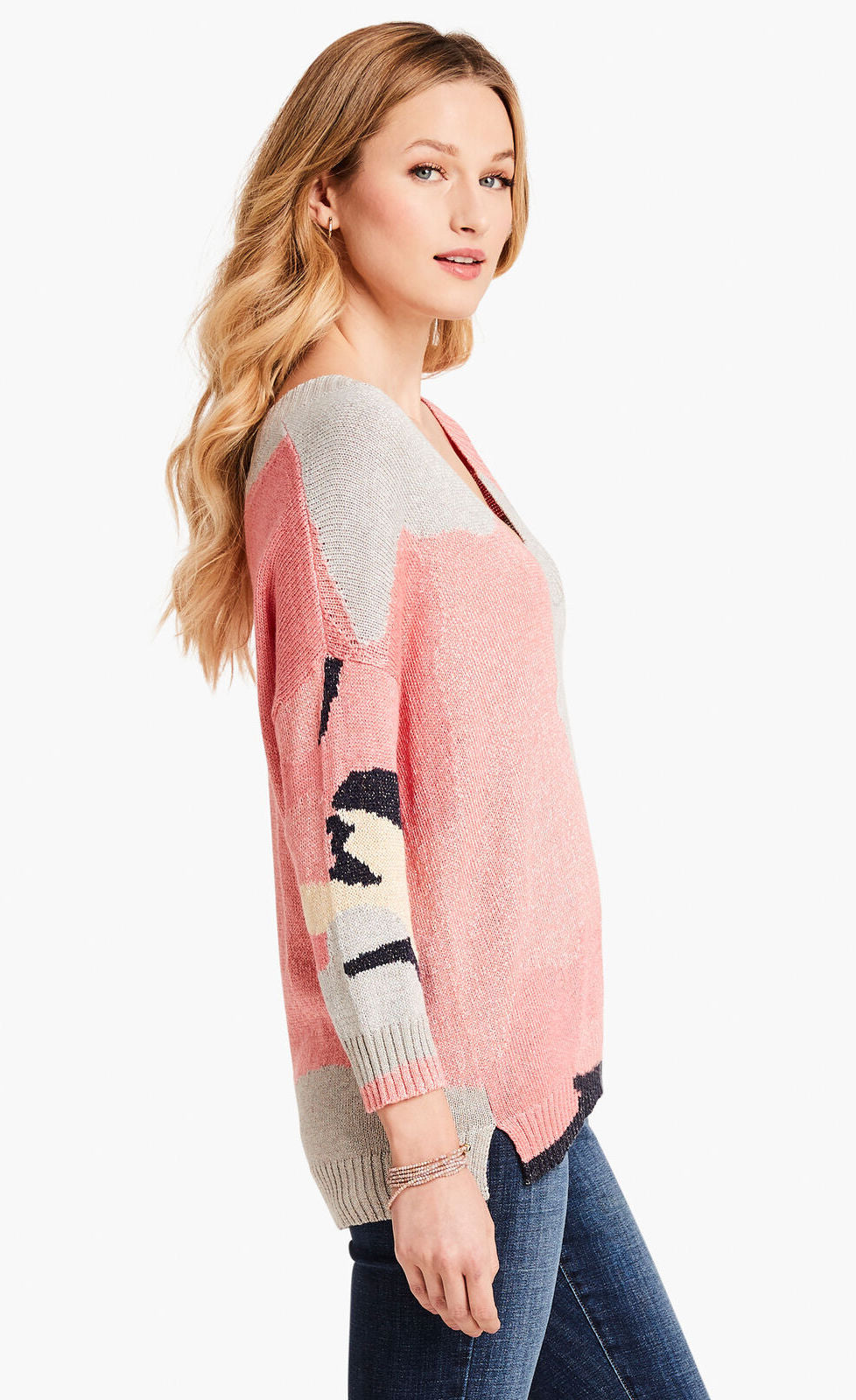 Right-sided top half view of a model wearing the nic+zoe citrus splash sweater. This sweater is mainly pink with abstract colors of grey, blue, and creme all over it. This sweater has long drop shoulder sleeves.