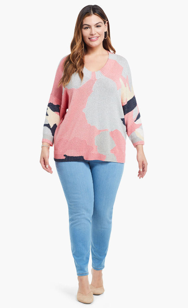 Front full body view of a model wearing the nic+zoe citrus splash sweater in a plus size. This sweater is mainly pink with abstract colors of grey, blue, and creme all over it. This sweater has long drop shoulder sleeves.
