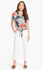Load image into Gallery viewer, Front full body view of a woman wearing white pants and the nic+zoe fan dot tie top. This top has a tie front, a v-neck, short sleeves, and a white background with navy dots and fanned navy, red, and nude flowers all over it.
