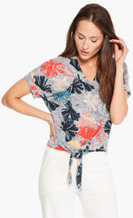 Load image into Gallery viewer, Front top half view of a woman wearing white pants and the nic+zoe fan dot tie top. This top has a tie front, a v-neck, short sleeves, and a white background with navy dots and fanned navy, red, and nude flowers all over it.
