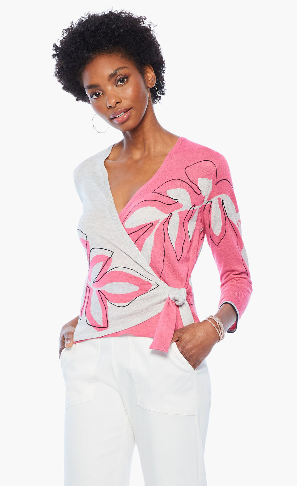 Front top half view of a woman wearing the nic+zoe hibiscus tie sweater. This sweater has a wrap look with a faux side tie. The left side of it is pink with a hibiscus flower while the right side is white with an outlined hibiscus flower. The sweater has 3/4 length sleeves.