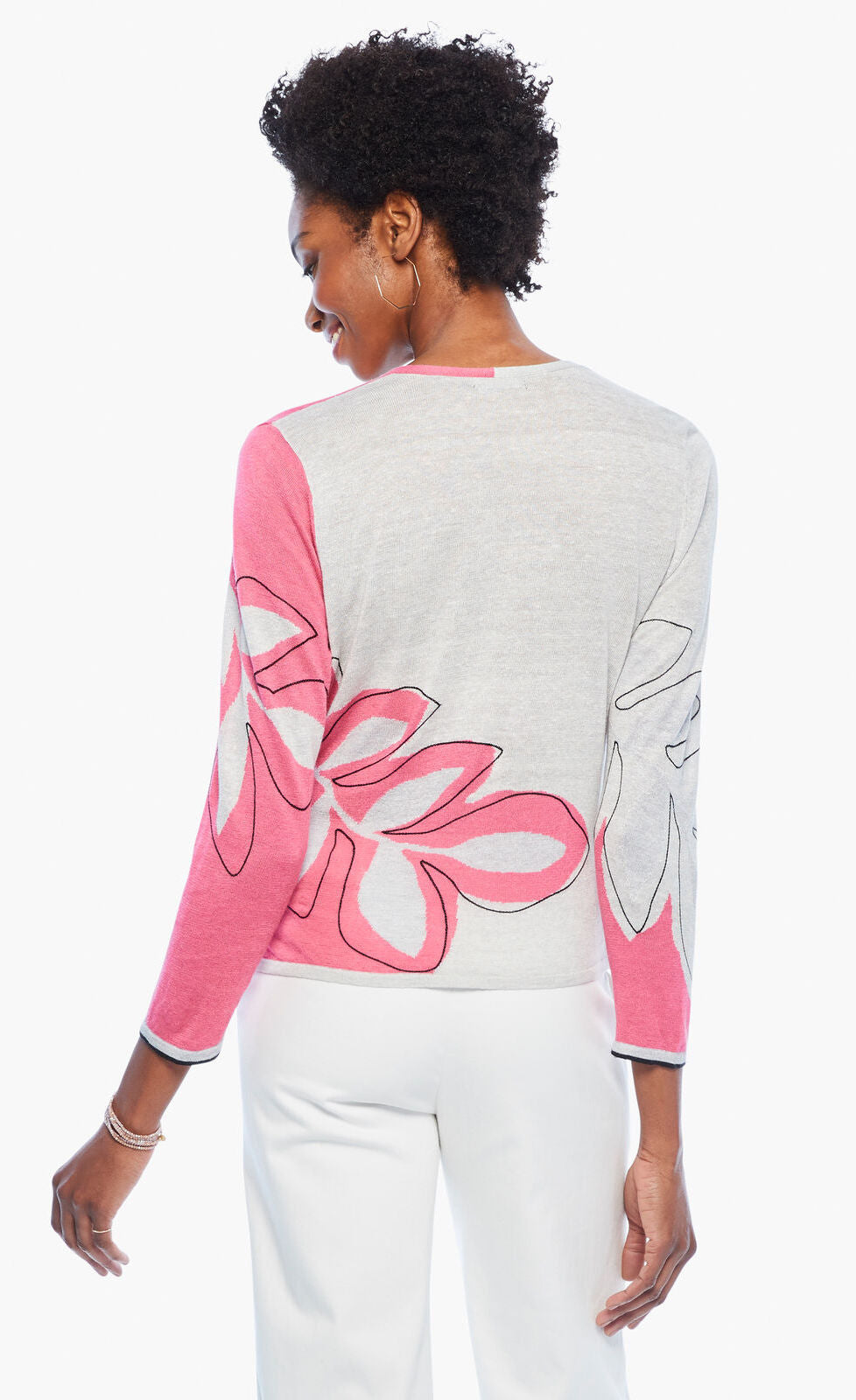 Back top half view of a woman wearing the nic+zoe hibiscus tie sweater. This sweater has a wrap look with a faux side tie. The back is white with a pink hibiscus flower while the sleeves are pink and white. The sweater has 3/4 length sleeves.