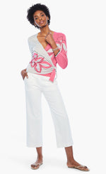 Load image into Gallery viewer, Front full body view of a woman wearing white pants and the nic+zoe hibiscus tie sweater. This sweater has a wrap look with a faux side tie. The left side of it is pink with a hibiscus flower while the right side is white with an outlined hibiscus flower. The sweater has 3/4 length sleeves.

