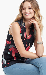 Front view of woman sitting and wearing light blue jeans and the kaleidoscope tank top from Nic+Zoe. The tank is black with abstract pink and indigo print. 