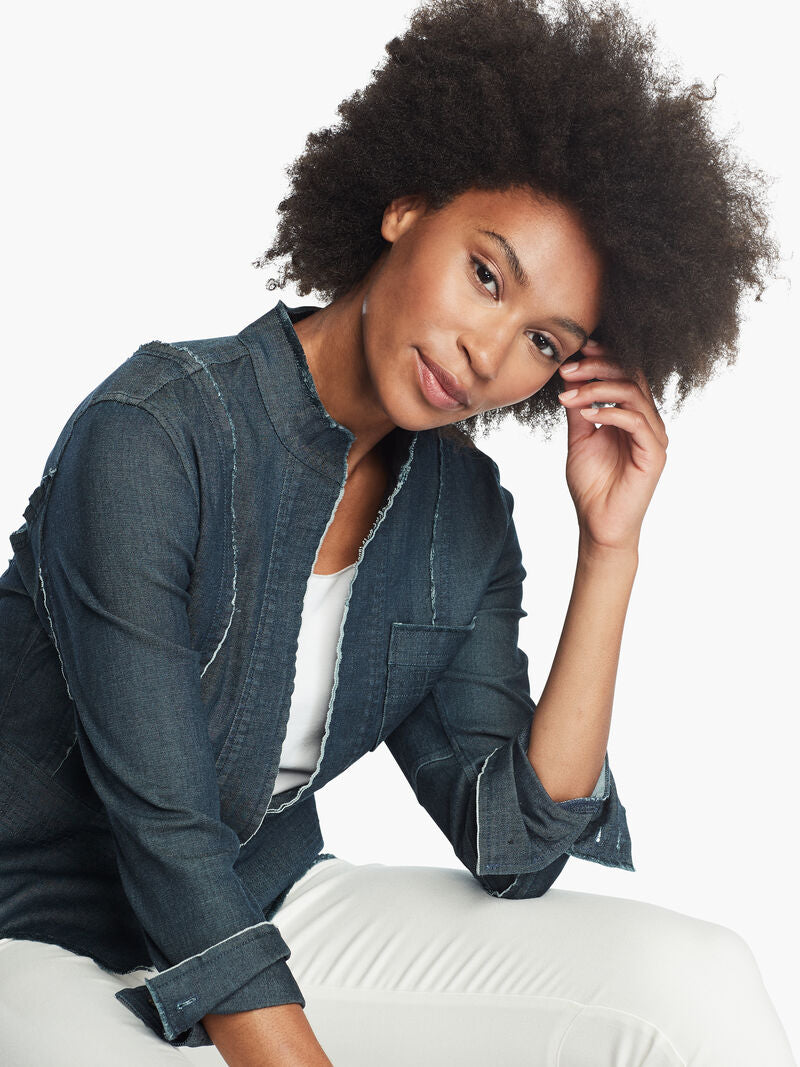 Front, top half view of a woman sitting leaned forward wearing white pants and the Nic + Zoe Favorite Denim Jacket. The dark denim jacket features frayed stitching, long sleeves that can be cuffed, and a short stand collar.