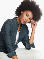 Load image into Gallery viewer, Front, top half view of a woman sitting leaned forward wearing white pants and the Nic + Zoe Favorite Denim Jacket. The dark denim jacket features frayed stitching, long sleeves that can be cuffed, and a short stand collar.
