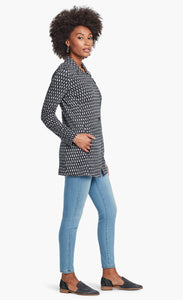 Right side, full body view of a woman with her hands in the front pocket of the Nic + Zoe Ponte Ikat Jacket. On the bottom she is wearing fitted jeans. The closed jacket is indigo with a white speck print all over it. It has a flat, notched lapel, and a front, off-center zipper.