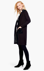 Load image into Gallery viewer, Left side full body view of a woman wearing the nic+zoe royal trimmings jacket and black leggings. This jacket is black with faux leather trim and two faux leather front pockets. The jacket has long sleeves and goes down to the knees.
