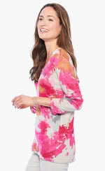 Load image into Gallery viewer, Left side top half view of a woman wearing white pants and the nic+zoe sunset blooms sweater. This sweater has a v-neck and 3/4 length sleeves. It&#39;s white based with pink, orange, and grey splashes of color all over it.
