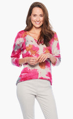 Load image into Gallery viewer, Front top half view of a woman wearing white pants and the nic+zoe sunset blooms sweater. This sweater has a v-neck and 3/4 length sleeves. It&#39;s white based with pink, orange, and grey splashes of color all over it.
