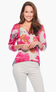 Front top half view of a woman wearing white pants and the nic+zoe sunset blooms sweater. This sweater has a v-neck and 3/4 length sleeves. It's white based with pink, orange, and grey splashes of color all over it.