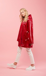 Load image into Gallery viewer, Left side full body view of a woman with blonde hair wearing white pants, white shoes, and the red Courtney Raincoat from Nikki Jones. This raincoat has long sleeves, a hood, snakeskin print, and comes down below the hips
