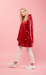 Left side full body view of a woman with blonde hair wearing white pants, white shoes, and the red Courtney Raincoat from Nikki Jones. This raincoat has long sleeves, a hood, snakeskin print, and comes down below the hips