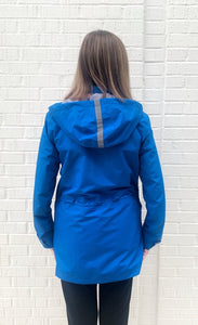 Back top half view of a woman wearing the nikki jones royal blue magic print rain jacket. This jacket has a hood and adjustable waist. It sits below the hips.