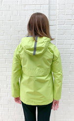 Load image into Gallery viewer, Back top half view of woman wearing the nikki jones magic print lime rain jacket. The back of this jacket has a hood and venting.
