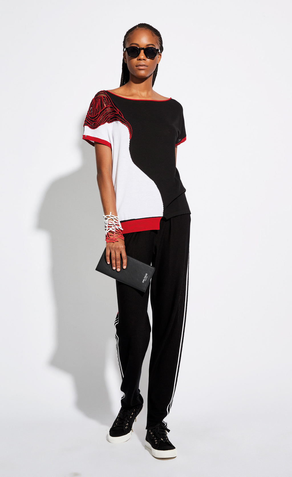 Front full body view of a woman wearing sunglasses, black sweat pants, and the Oblique Creations Knit Tee. This tee is a mix of black, red, and white, with black and red cut out detailing on the right shoulder. 