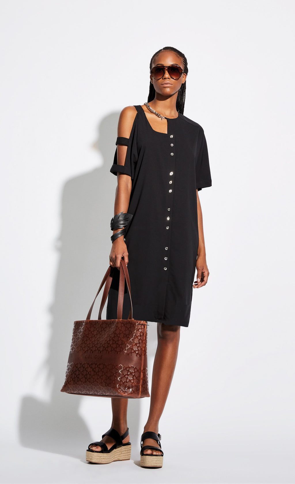 Front full body view of a woman wearing the Oblique Creations Black Cutout Dress. This dress has a button down front with every three buttons grouped off. The dress goes down to the model's knees. The right sleeve has cutouts to expose the arm. The right portion of the neckline is cut out and has a silver chain around it. The model is also holding a large brown hand bag and wearing large brown sunglasses.